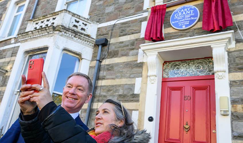 A woman holds a phone to take a selfie next to a man, in front of a house which has a blue plaque on the wall.
