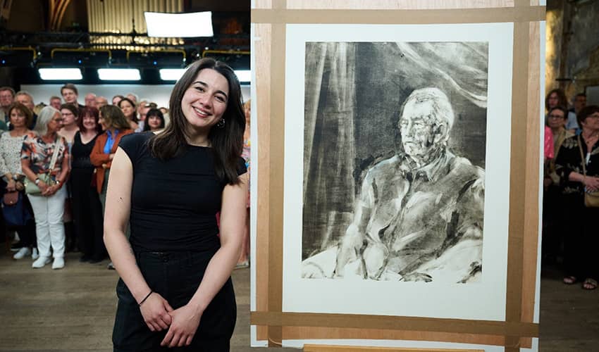 A woman wearing a black outfit stands beside a large portrait, with a crowd of people gathered in the background
