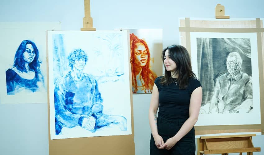 A woman in a black outfit stands in front of four large portrait artworks.