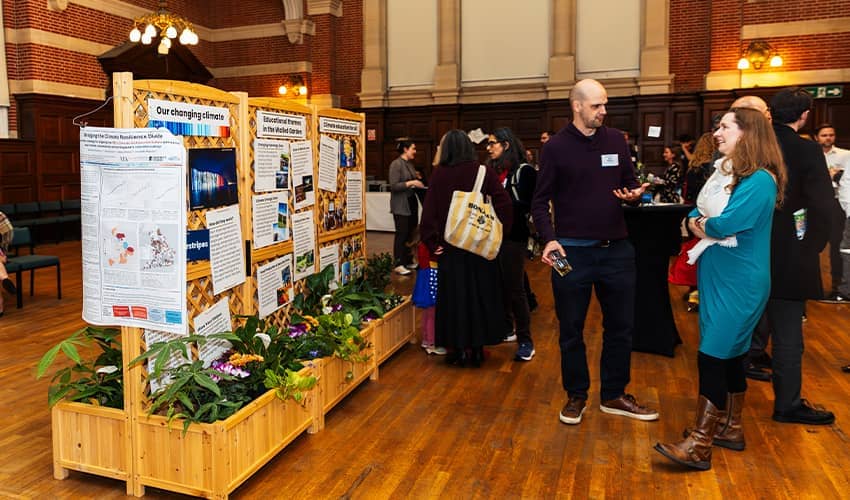 A group of people stood in a large hall speaking to each other next to some large information poster boards with images and presentation slides 