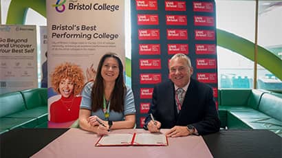 A woman and a man wearing business attire sit at a table with a document in front of them, they have pens in their hands to sign the document. They are sat in front of branded business banners and smiling at the camera.. 