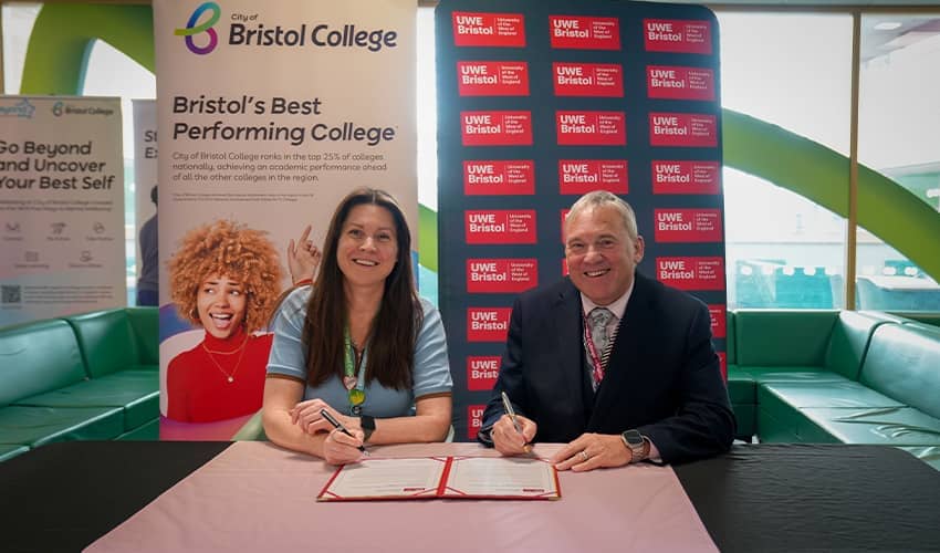 A woman and a man wearing business attire sit at a table with a document in front of them, they have pens in their hands to sign the document. They are sat in front of branded business banners and smiling at the camera.