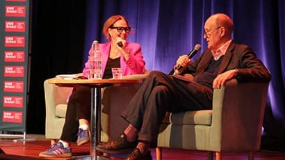 Two speakers sit onstage in armchairs, talking into microphones during a panel discussion.