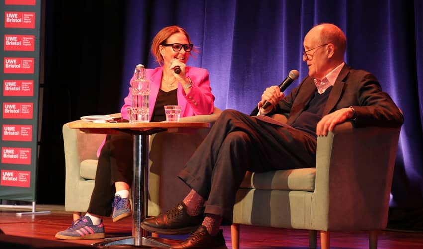 Two speakers sit onstage in armchairs, talking into microphones during a panel discussion.