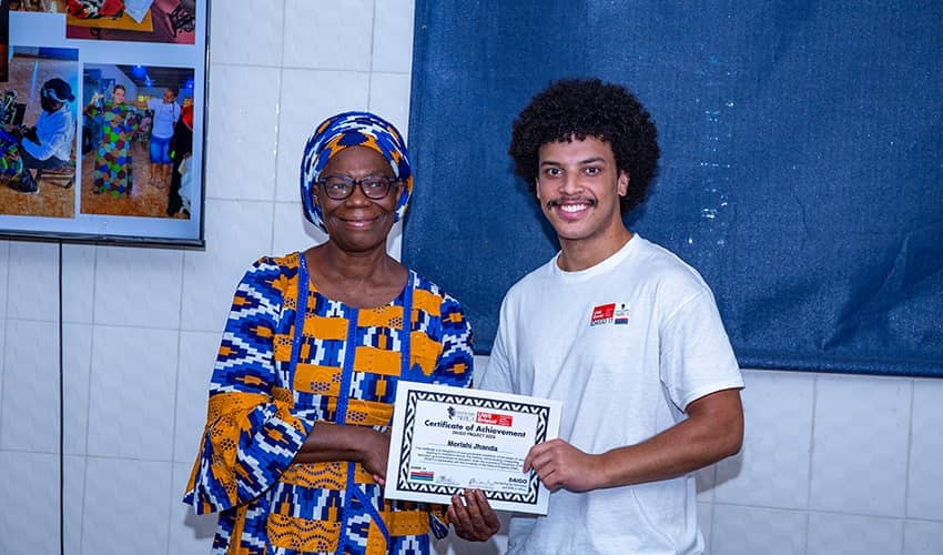 A woman and a man smile as they shake hands and hold a certificate between them. The woman is wearing traditional African clothing and the man is wearing a white tshirt with a UWE Bristol logo. 