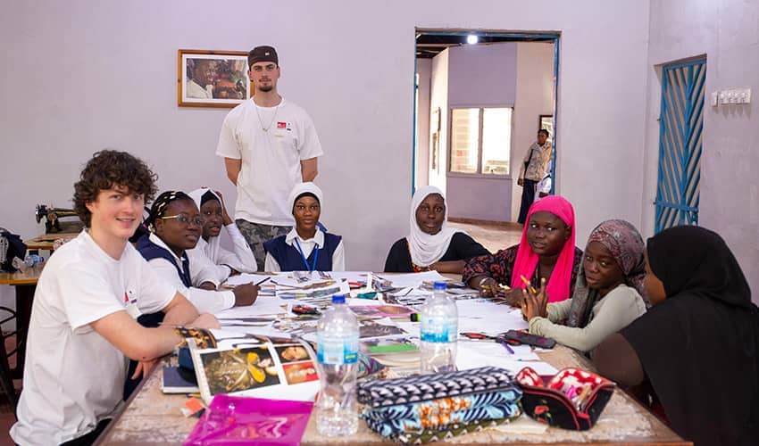 A group of people sat around a large table which is covered with books, pencil cases and fabric.