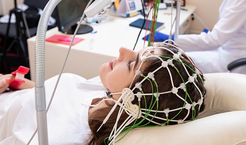 A woman is lying down in a hospital with a series of wires covering her head to monitor brain activity.