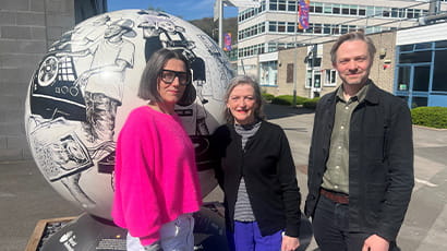 Two women and a man stand looking at the camera, with buildings and a globe sculpture behind them.
