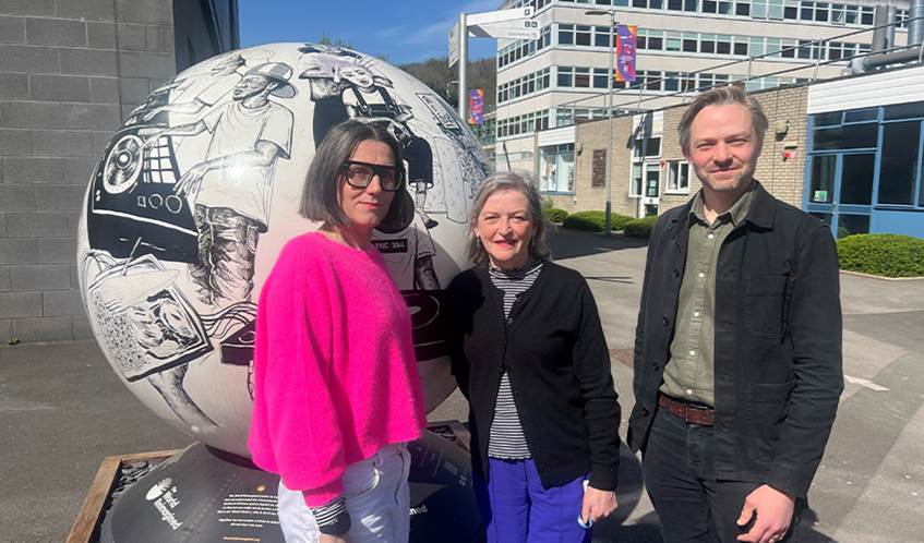 Two women and a man stand looking at the camera, with buildings and a globe sculpture behind them.