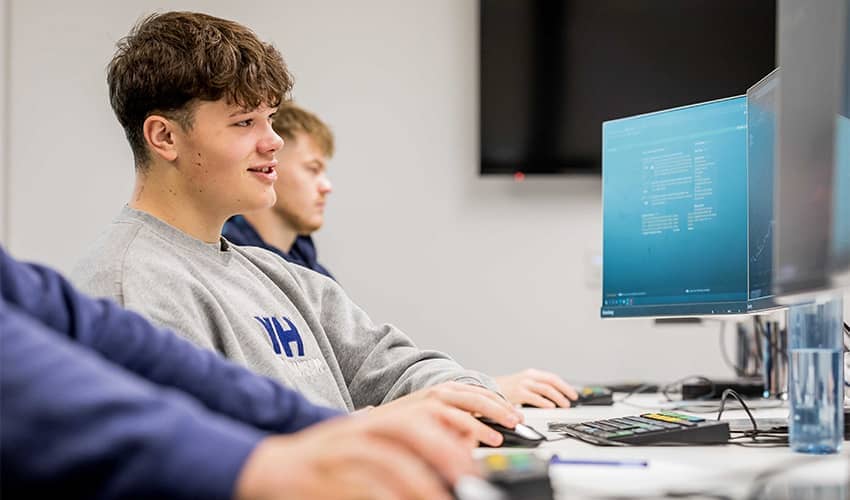 Three young male adults sit in front of computer screens in a seminar room.
