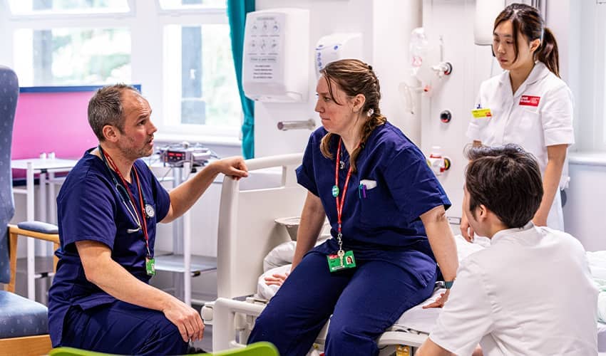 Two people (lecturers) wearing blue scrubs look at each other. One is kneeling the other sitting on  hospital bed. Two other people (students) wearing white uniforms look on listening. The background shows a window, chairs and medical equipment..