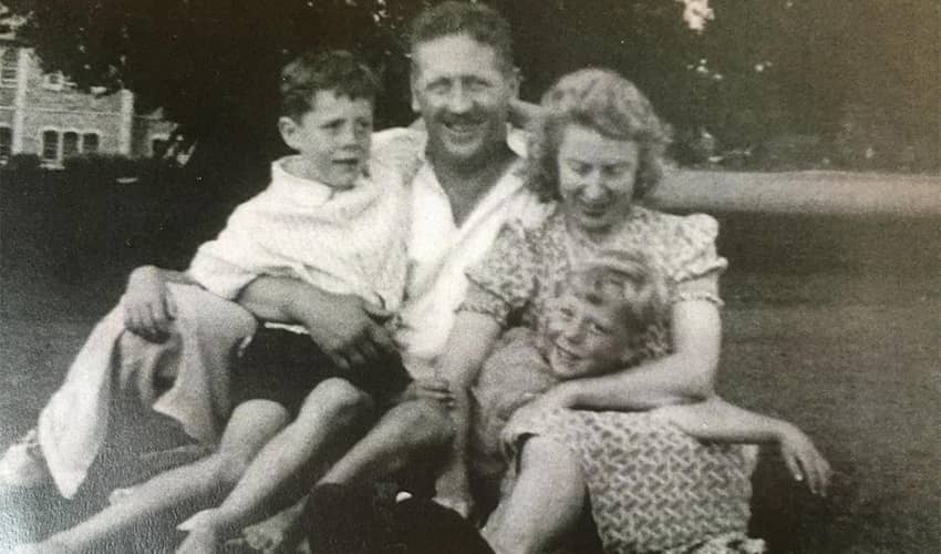 A black and white photo showing two adults and two children sitting on grass in front of trees and building.