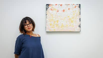 A woman standing next to a painting which is hung on a plain white wall. The painting is abstract which white, cream, orange and blue colours. 