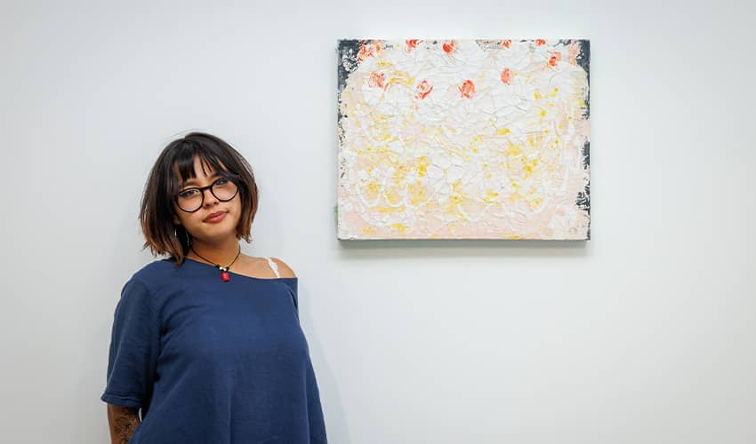 A woman standing next to a painting which is hung on a plain white wall. The painting is abstract which white, cream, orange and blue colours. 