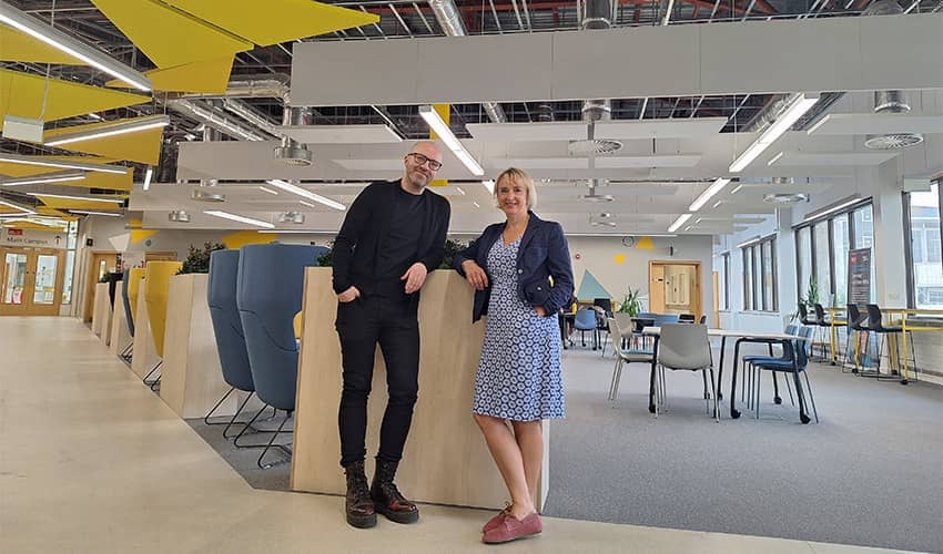 A man and a woman stand in a large reception area of a business with industrial exposed pipework in the ceiling and yellow triangles hanging from the ceiling, and workspaces in the background. They are smiling at the camera.