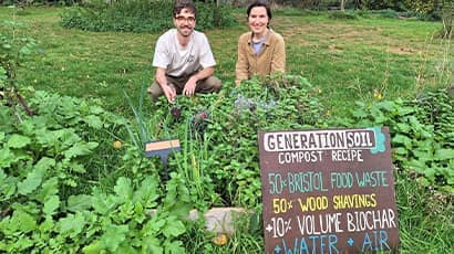 Two people crouch behind green shubbery in a garden with a large sign propped up at the front of the image saying 'Generation Soil Compost Recipe' 