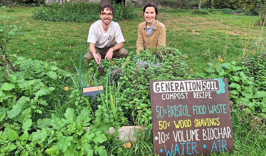 Two people crouch behind green shubbery in a garden with a large sign propped up at the front of the image saying 'Generation Soil Compost Recipe' 
