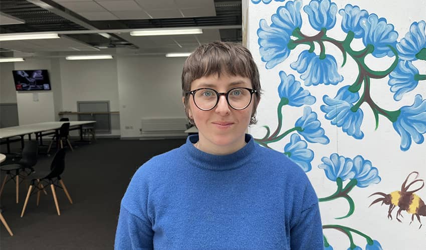 A person in classroom is smiling at a camera wearing glasses and a blue jumper. The person is standing in front of a white wall which has blue flowers and bumblebee decoration. 