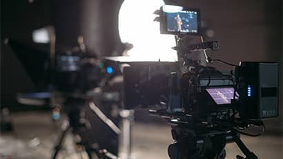 A close up shot of a large film camera on a tripod facing a studio setting which is blurred in the background. 