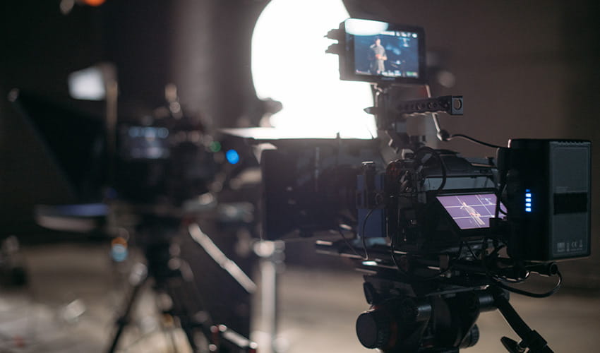 A close up shot of a large film camera on a tripod facing a studio setting which is blurred in the background. 