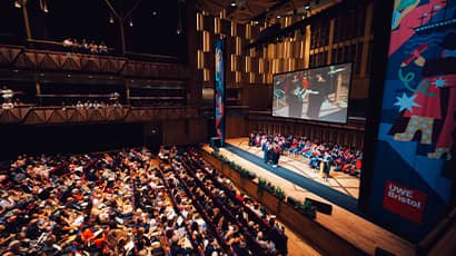 A large auditorium filled with an audience watching a stage with a graduation ceremony taking place and a student in a cap and gown walking across the stage.