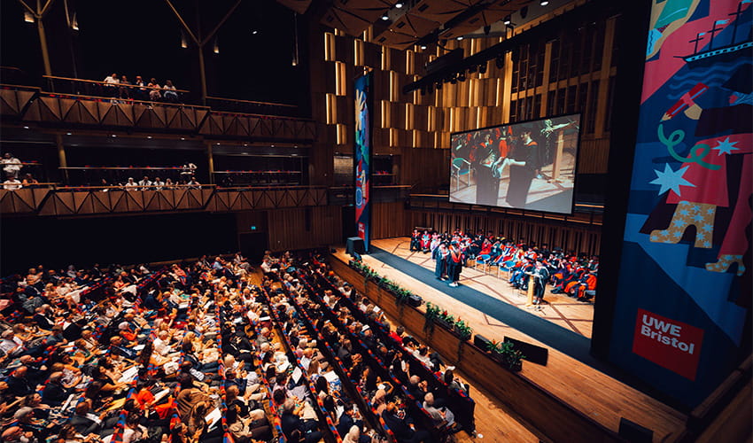 A large auditorium filled with an audience watching a stage with a graduation ceremony taking place and a student in a cap and gown walking across the stage.