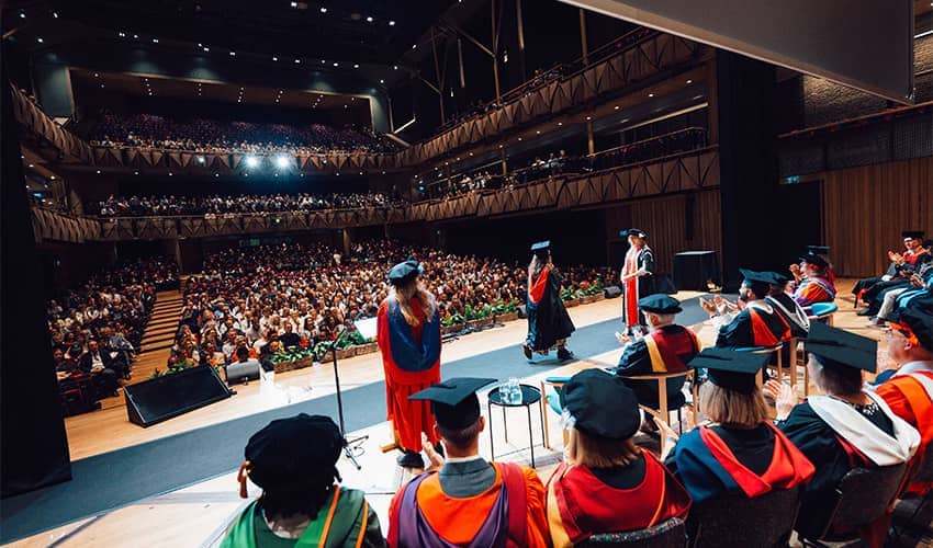 A wide angle shot of a large auditorium with hundreds of people sat facing a stage which has individuals dressed in cap and gowns walking across the stage. 