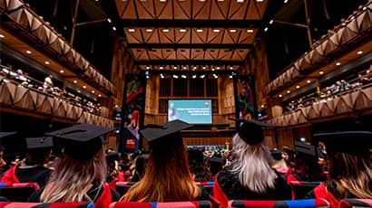Four women sit in a large auditorium in a university graduation ceremony with their back to the camera wearing a mortar board and gown 