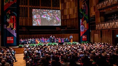 A large theatre with a full audience and a person crossing a stage wearing a graduation cap and gown shaking the hand of another person.