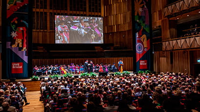 A large theatre with a full audience and a person crossing a stage wearing a graduation cap and gown shaking the hand of another person.
