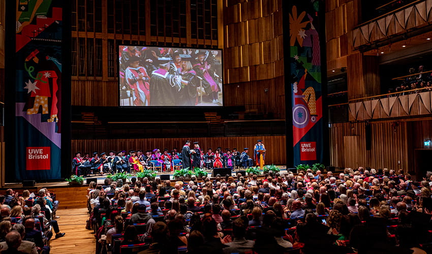 A large theatre auditorium with a full audience and a person crossing a stage wearing a graduation cap and gown shaking the hand of another person.
