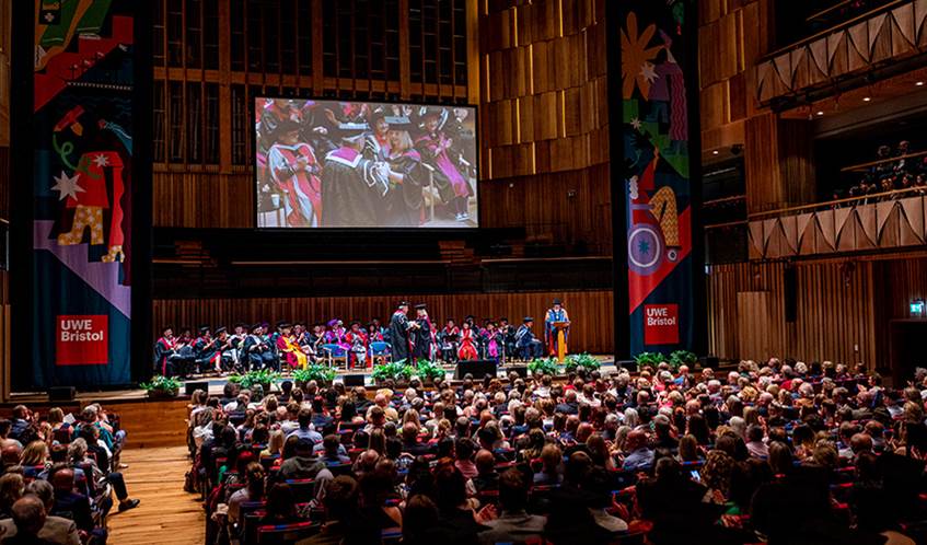 A large theatre auditorium with a full audience and a person crossing a stage wearing a graduation cap and gown shaking the hand of another person.