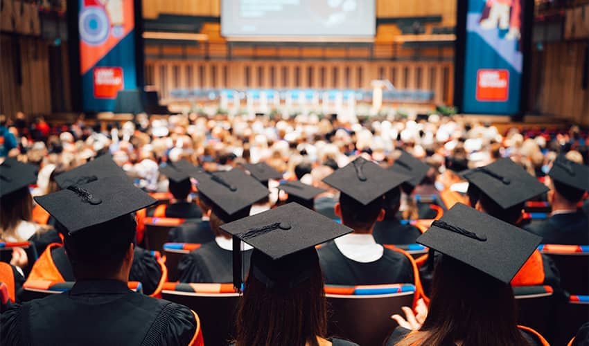 The backs of heads of hundreds of graduates sat in an auditorium wearing cap and gowns. 
