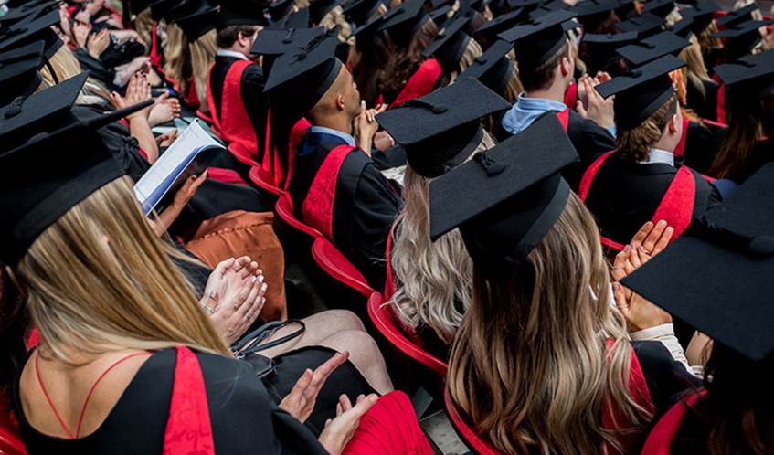 Students wearing graduation robes and mortarboard hats sit clapping as someone receives their degree