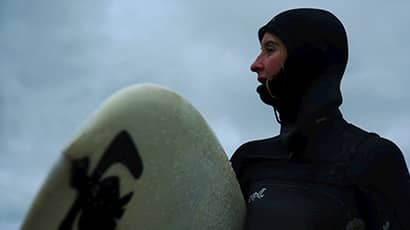 A head and shoulders shot of a person wearing a wet suit and holding a surfboard. 