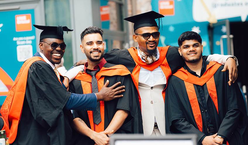 A group of four graduates wearing black gowns with orange stoles and mortarboards are standing together, posing for a photo.