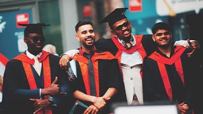 A group of four graduates wearing black caps and gowns with red stoles stand closely together, posing for a photo.