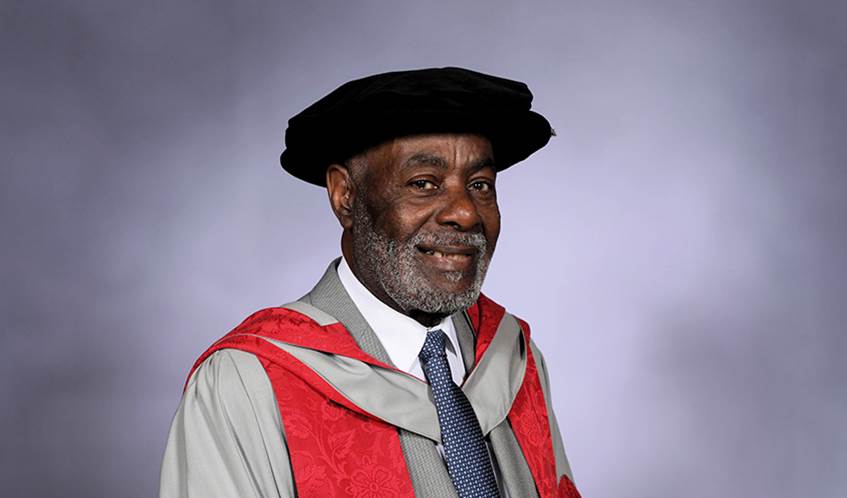 A man wearing a university hat and gown smiles against a grey background.