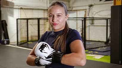 A girl wearing boxing gloves standing in a gym smiles at the camera