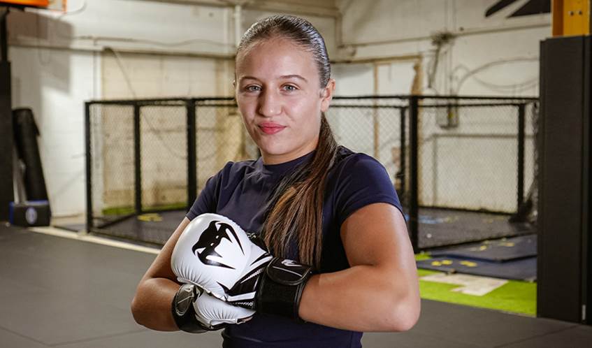 A girl wearing boxing gloves standing in a gym smiles at the camera