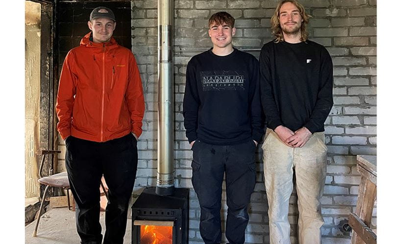 Three people stood next to a brick wall with a wood burning stove between them that is lit
