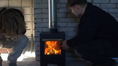 Two people crouch in front of a wood burning stove 