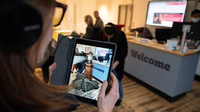 A woman stands in an office looking at a mobile phone screen which shows a virtual reality image of a lighthouse standing on rocks. 