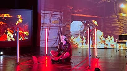 A person sits on a stage floor with gaming equipment, surrounded by vertical bright lights, in front of large projected images of afire scene.