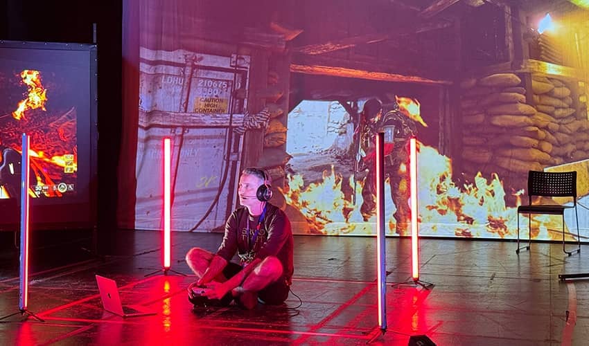 A person sits on a stage floor with gaming equipment, surrounded by vertical bright lights, in front of large projected images of a fire scene.