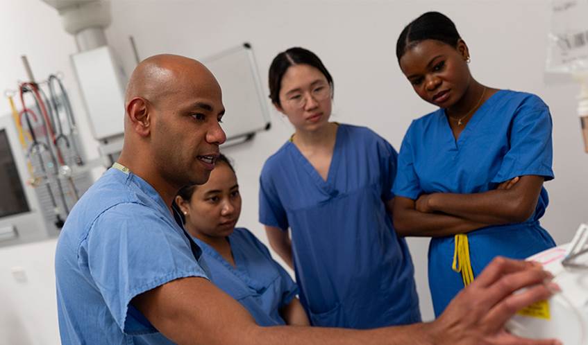 Four people wearing hospital scrubs in a healthcare setting gather around a piece of hospital equipment.