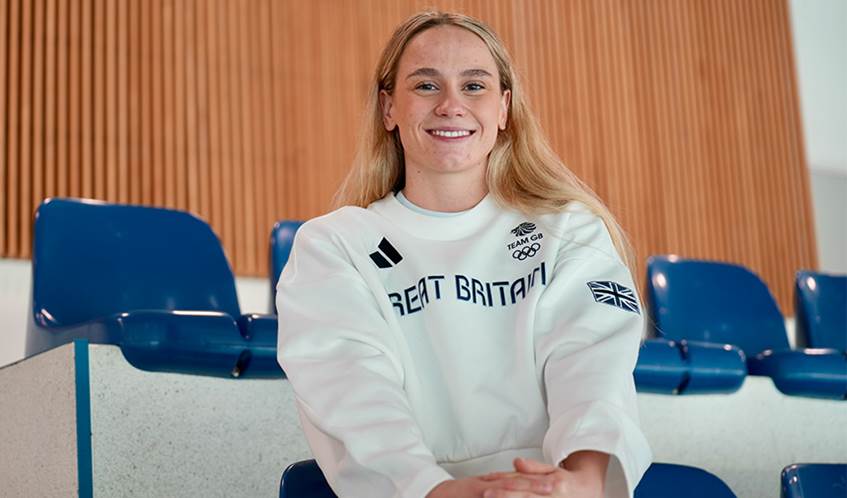 A woman wearing a Great Britain team sweatshirt smiles whilst sitting in spectator seats at a swimming pool.