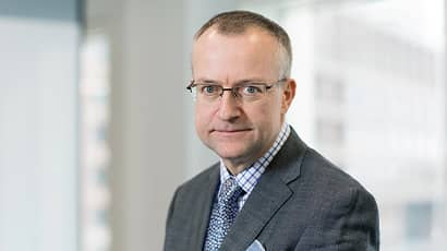 A head and shoulders image of a man wearing a suit with glasses looking down the lens of the camera in a corporate setting. 