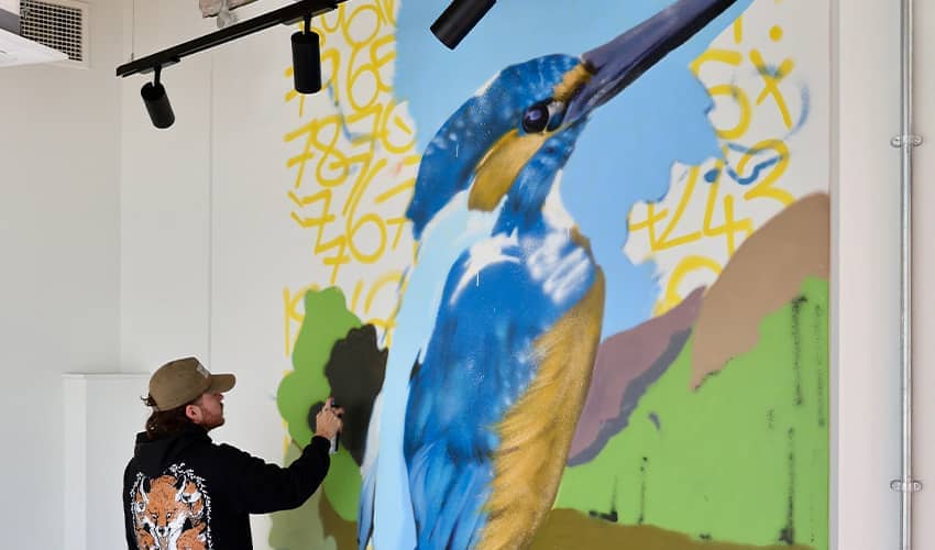 An artist paints a large scale mural of a colourful Kingfisher on a wall