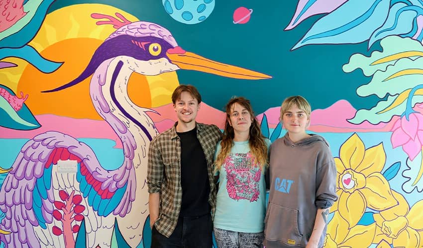 Three people stand in front of a painted colourful wildlife mural